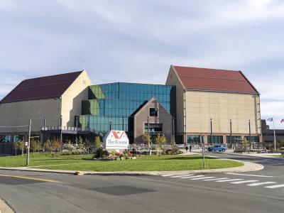 A large modern cultural building with two prominent gabled wings and a central glass atrium, featuring a white sign in the foreground lawn that reads "The Rooms".