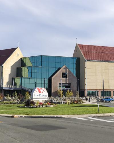 A large modern cultural building with two prominent gabled wings and a central glass atrium, featuring a white sign in the foreground lawn that reads "The Rooms".