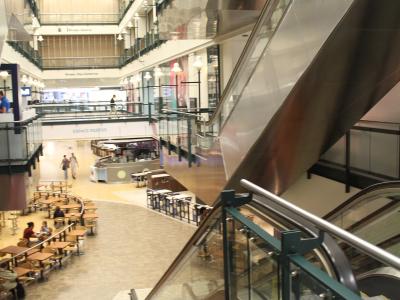 A multi-level indoor shopping complex featuring a large atrium with a grid-patterned skylight, escalators, and a food court area labeled "ESPACE RESTOS".