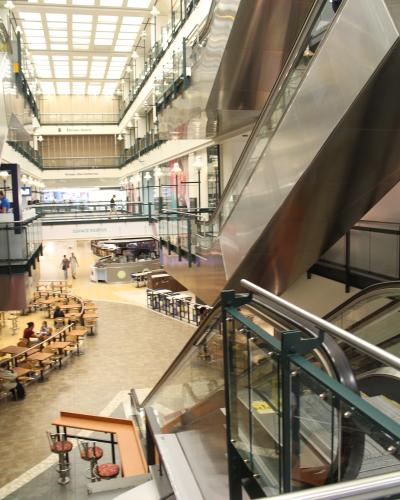 A multi-level indoor shopping complex featuring a large atrium with a grid-patterned skylight, escalators, and a food court area labeled "ESPACE RESTOS".