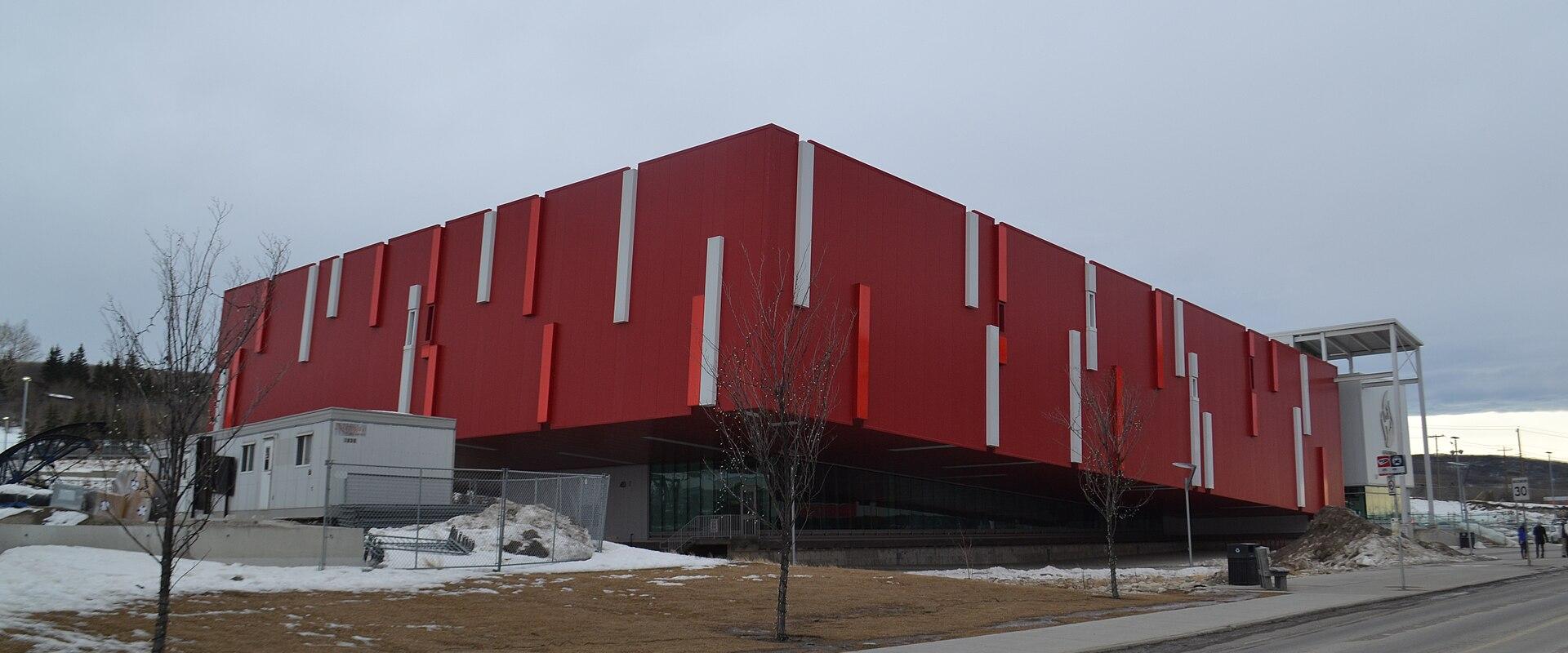 A large, modern building with a distinctive red facade featuring vertical white and orange accents, situated in a snowy, overcast environment.