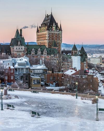 A panoramic winter view of the Quebec City skyline featuring the prominent Château Frontenac hotel, historic stone buildings, and a snow-covered park in the foreground overlooking the St. Lawrence River.