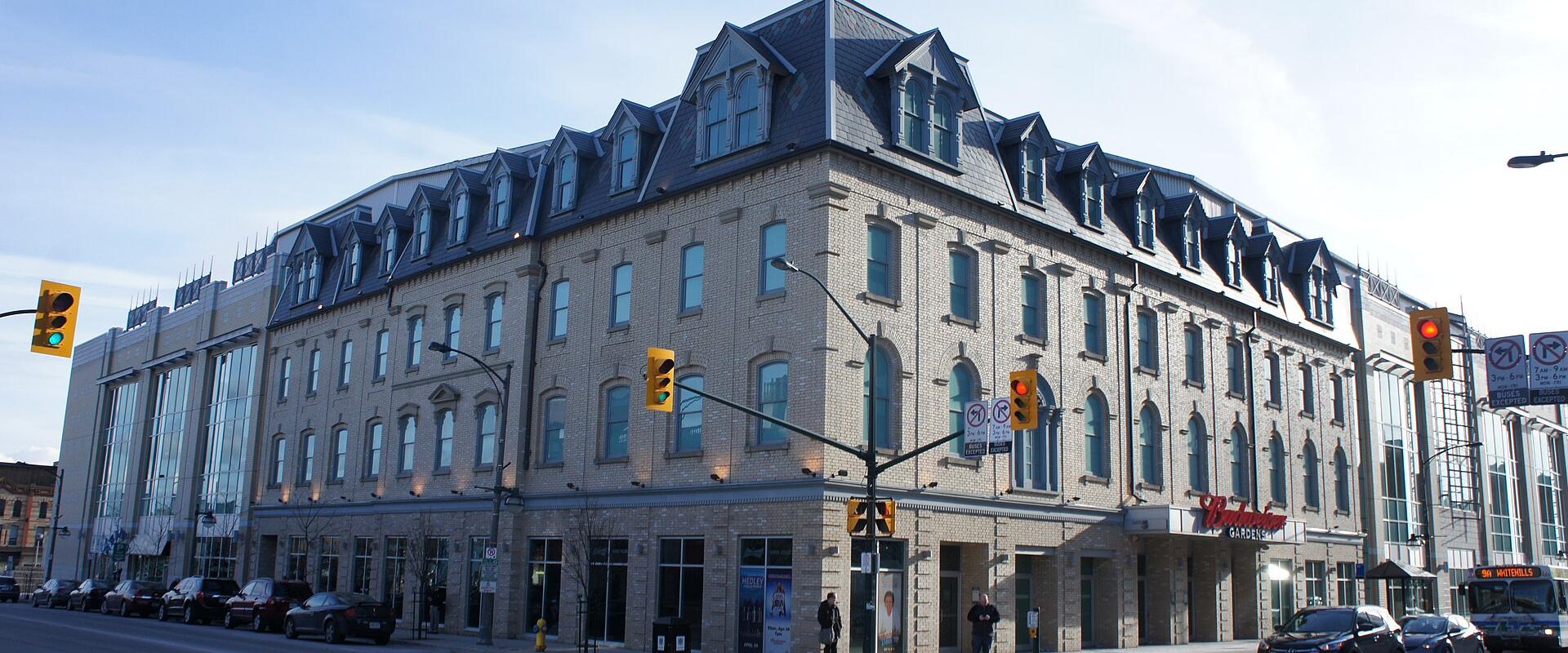 A large, multi-story brick building with a mansard roof and dormer windows situated on a city street corner, featuring a red "Budweiser Gardens" sign above the entrance and a city bus on the right.