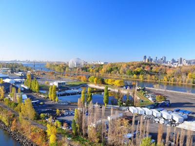 A high-angle panoramic view of Parc Jean-Drapeau on a sunny autumn day, featuring the Montreal Biosphere, the Olympic Stadium in the distance, and the Montreal skyline across the river.