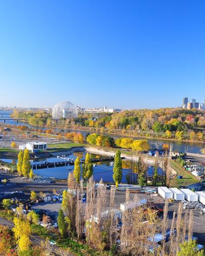 A high-angle panoramic view of Parc Jean-Drapeau on a sunny autumn day, featuring the Montreal Biosphere, the Olympic Stadium in the distance, and the Montreal skyline across the river.