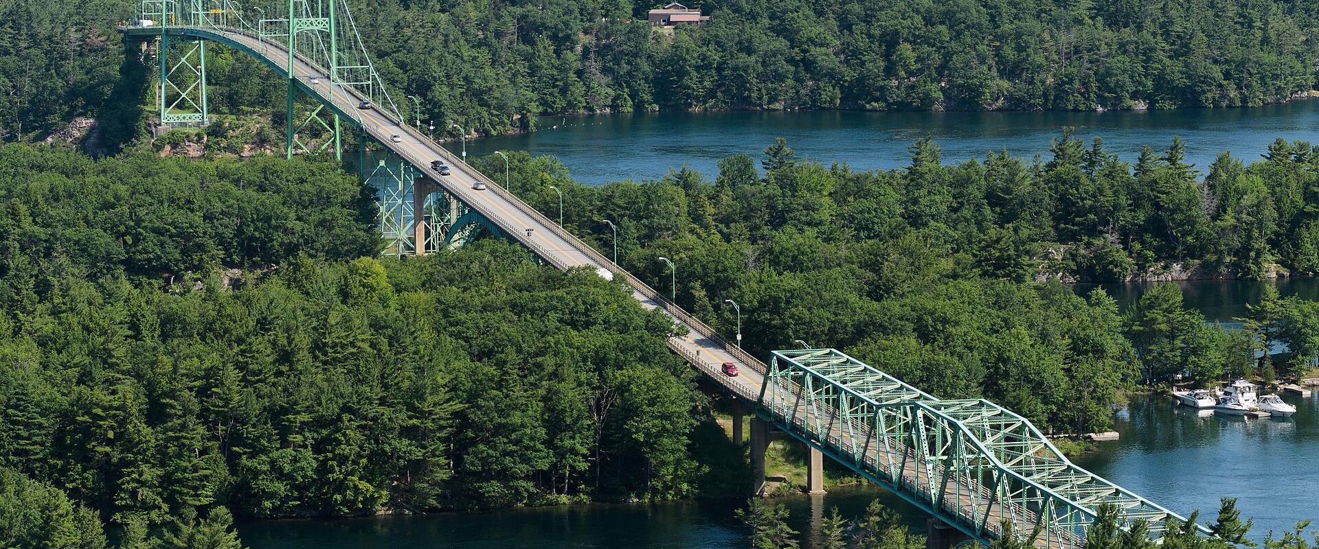 A high-angle view of a long, green-painted steel truss bridge system spanning a river channel between densely forested islands, featuring a distinct vertical-lift span section and a continuous truss approach.