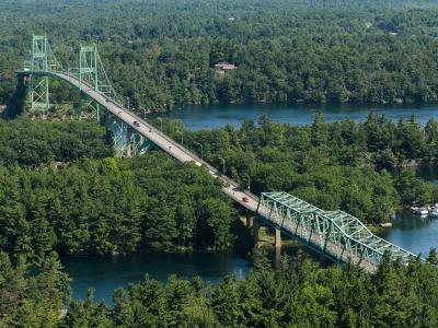 A high-angle view of a long, green-painted steel truss bridge system spanning a river channel between densely forested islands, featuring a distinct vertical-lift span section and a continuous truss approach.