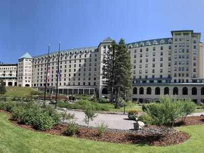 A large, multi-winged historic hotel with a distinctive green copper roof and beige stone facade, set against a backdrop of evergreen trees and a clear blue sky, with a Canadian flag flying in the foreground.