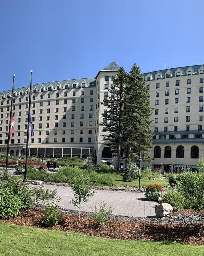 A large, multi-winged historic hotel with a distinctive green copper roof and beige stone facade, set against a backdrop of evergreen trees and a clear blue sky, with a Canadian flag flying in the foreground.