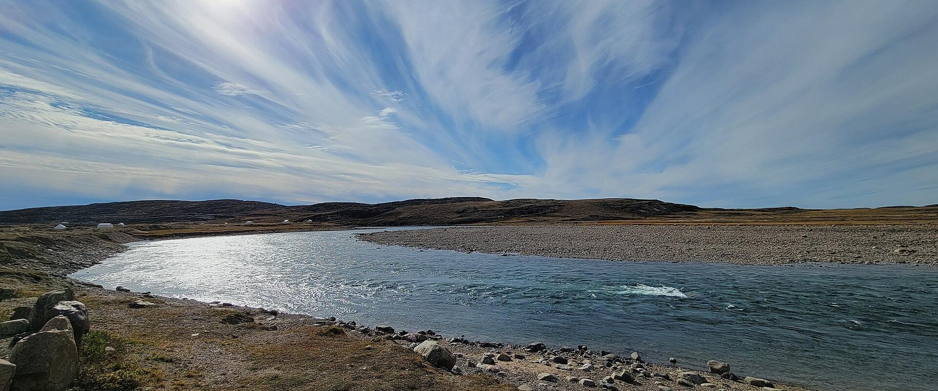 A wide-angle landscape photograph showing a river flowing through a barren, rocky tundra landscape under a bright sun and streaked clouds, with small white structures visible on the distant shore.