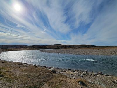 A wide-angle landscape photograph showing a river flowing through a barren, rocky tundra landscape under a bright sun and streaked clouds, with small white structures visible on the distant shore.