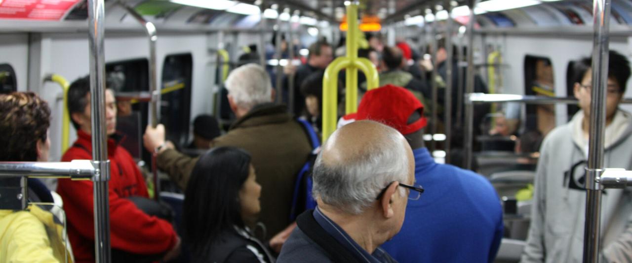 Interior of a crowded automated light rail train car with passengers standing and holding onto vertical metal poles and yellow handrails.