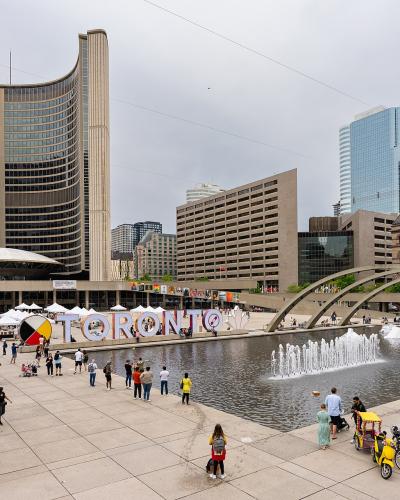 A wide shot of a public plaza featuring the distinctive curved Toronto City Hall buildings, a reflecting pool with fountains, and large white letters spelling "TORONTO" with a red maple leaf logo.