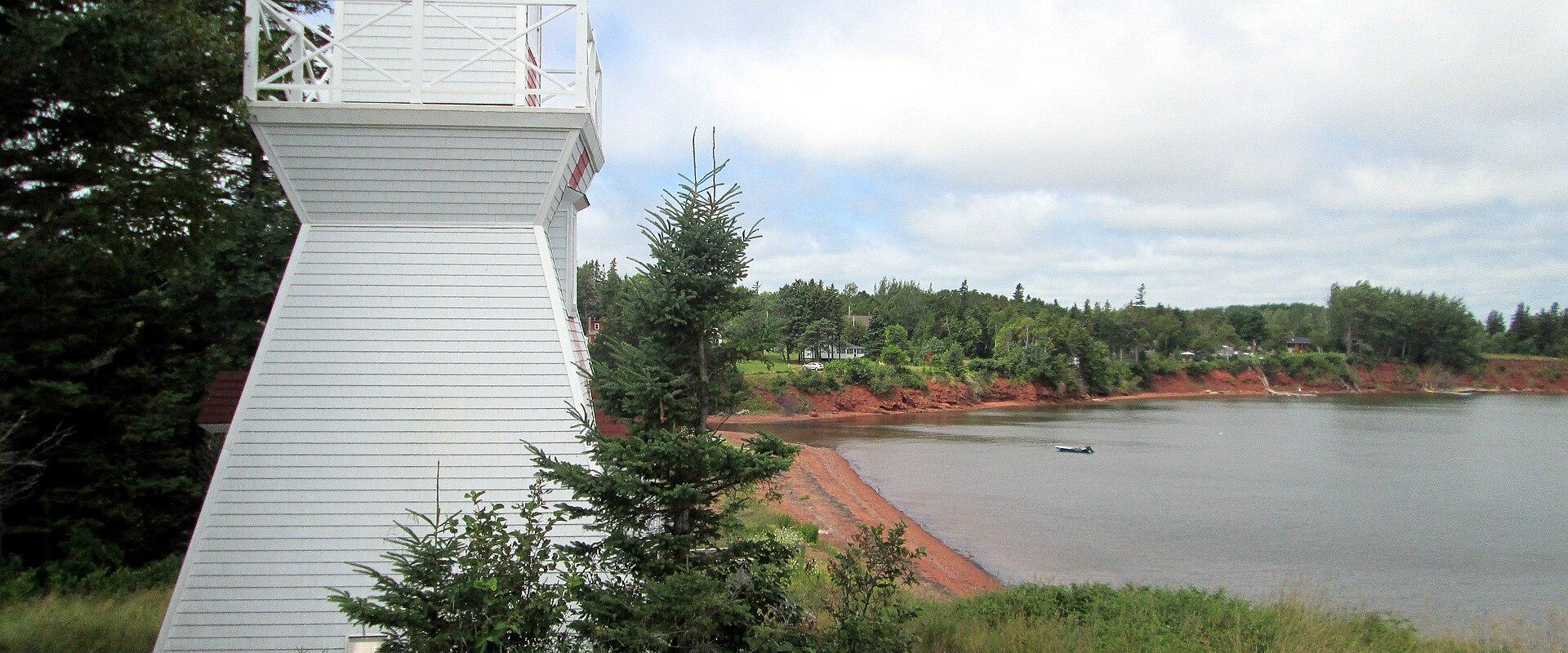 A white wooden lighthouse with a red roof stands on a grassy slope next to a body of water with red soil banks and a forested shoreline in the background.