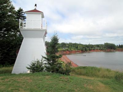 A white wooden lighthouse with a red roof stands on a grassy slope next to a body of water with red soil banks and a forested shoreline in the background.