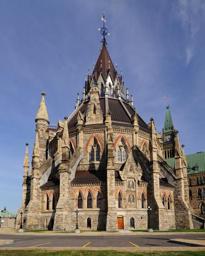 A low-angle photograph of the Library of Parliament, a Gothic Revival stone building with a central spire and flying buttresses, with the Peace Tower visible in the background.
