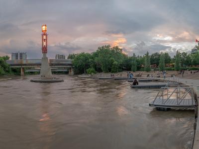 A wide shot of a river confluence at dusk featuring a distinctive red lighthouse structure on a concrete base, floating docks, and a bridge in the background.