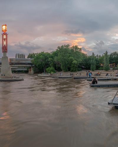 A wide shot of a river confluence at dusk featuring a distinctive red lighthouse structure on a concrete base, floating docks, and a bridge in the background.