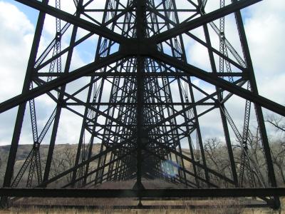 A low-angle view looking up through the massive steel truss structure of a railway bridge, showing the lattice of girders against a blue sky with clouds and dry grassy terrain below.