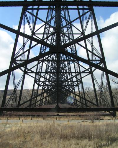 A low-angle view looking up through the massive steel truss structure of a railway bridge, showing the lattice of girders against a blue sky with clouds and dry grassy terrain below.