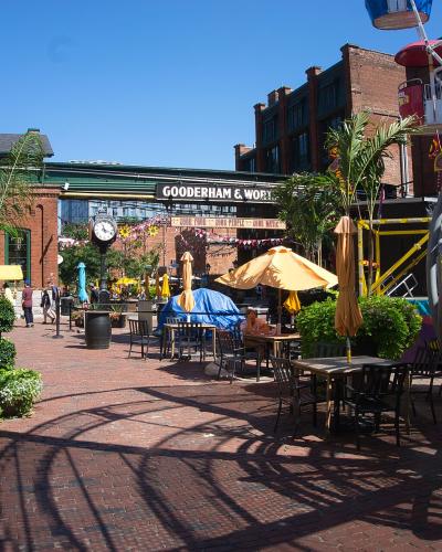 A brick-paved pedestrian plaza with outdoor dining tables, umbrellas, and a large metal sculpture in the foreground, featuring a prominent black sign reading "GOODERHAM & WORTS" on a brick building in the background.