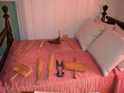 A bed with a red patterned coverlet and striped pillows, displaying a collection of antique wooden and metal textile-making tools (including shuttles, a carding brush, and a wiper) arranged on the mattress.