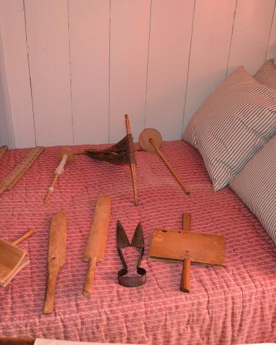 A bed with a red patterned coverlet and striped pillows, displaying a collection of antique wooden and metal textile-making tools (including shuttles, a carding brush, and a wiper) arranged on the mattress.