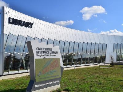 A modern white building with large glass windows and a sign reading "LIBRARY" on the facade, with a foreground sign identifying it as the "CIVIC CENTRE RESOURCE LIBRARY Vaughan Public Libraries" at "2191 Major Mackenzie Drive".