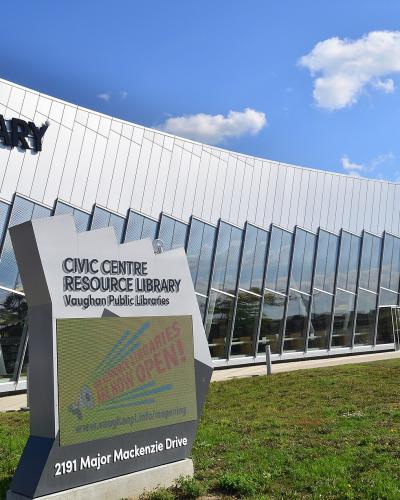 A modern white building with large glass windows and a sign reading "LIBRARY" on the facade, with a foreground sign identifying it as the "CIVIC CENTRE RESOURCE LIBRARY Vaughan Public Libraries" at "2191 Major Mackenzie Drive".