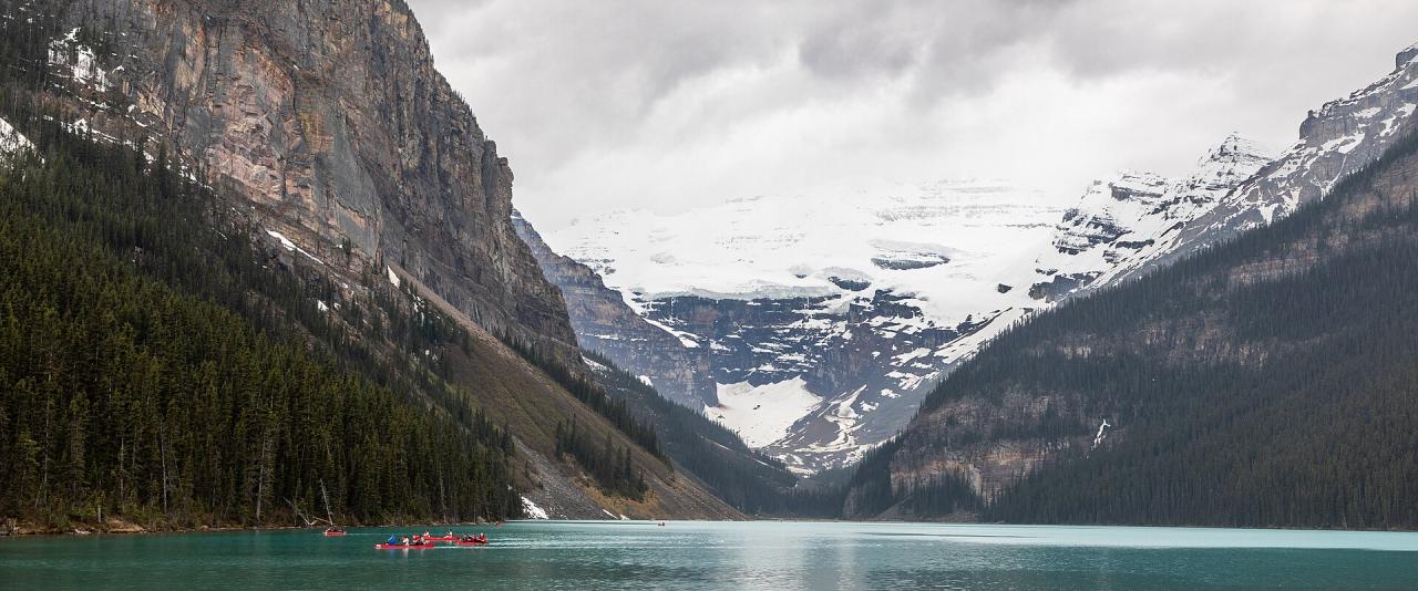 A wide shot of a turquoise alpine lake with red canoes on the water, flanked by steep, forested mountainsides and a large glacier in the background.