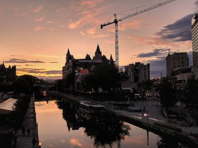 A sunset view of the Rideau Canal in Ottawa, featuring the distinctive châteauesque architecture of the Château Laurier hotel on the left bank and a construction crane in the background.