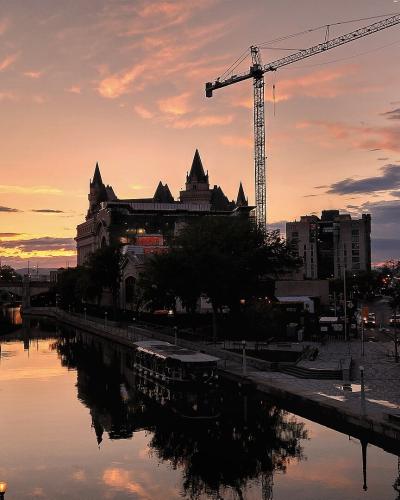 A sunset view of the Rideau Canal in Ottawa, featuring the distinctive châteauesque architecture of the Château Laurier hotel on the left bank and a construction crane in the background.
