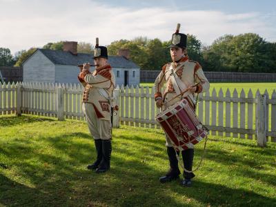 Two historical reenactors dressed in early 19th-century military uniforms, one playing a fife and the other a drum, standing on a grassy lawn in front of a white picket fence and a wooden building.