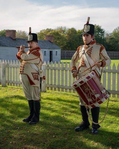Two historical reenactors dressed in early 19th-century military uniforms, one playing a fife and the other a drum, standing on a grassy lawn in front of a white picket fence and a wooden building.
