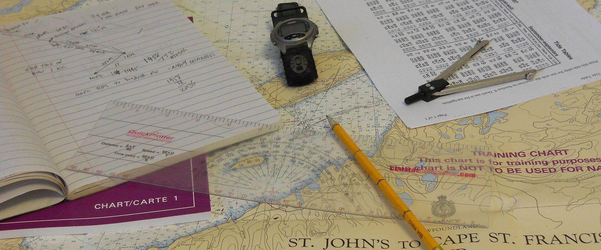 A close-up photograph of a nautical training chart, plotting tools, a notebook, and a watch laid out on a table.