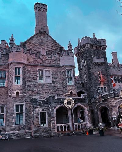 A low-angle photograph of a large, Gothic Revival stone castle featuring a prominent central tower, crenellated battlements, and holiday decorations including wreaths and a Canadian flag.