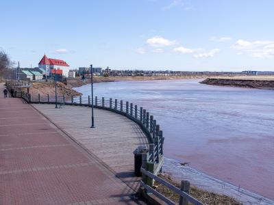 A paved and wooden boardwalk trail curving along the bank of a wide river with reddish-brown water, featuring a distinctive white building with a red roof on the left bank and a city skyline in the distance.