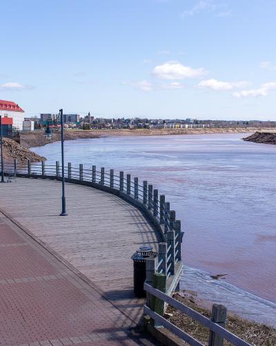 A paved and wooden boardwalk trail curving along the bank of a wide river with reddish-brown water, featuring a distinctive white building with a red roof on the left bank and a city skyline in the distance.