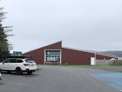 A white SUV is parked in front of a long, single-story building with reddish-brown siding and a distinctive angular roofline, with a green directional sign visible to the left.