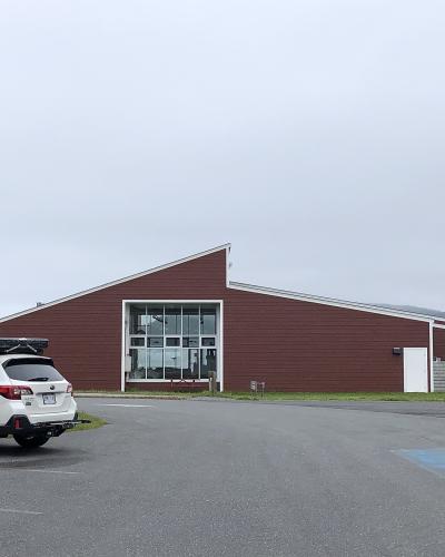 A white SUV is parked in front of a long, single-story building with reddish-brown siding and a distinctive angular roofline, with a green directional sign visible to the left.