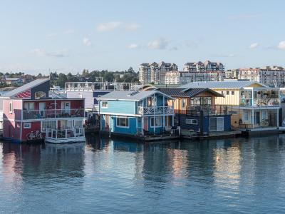 A row of colorful, multi-story houseboats moored in a harbor with modern apartment buildings visible on the shoreline in the background.