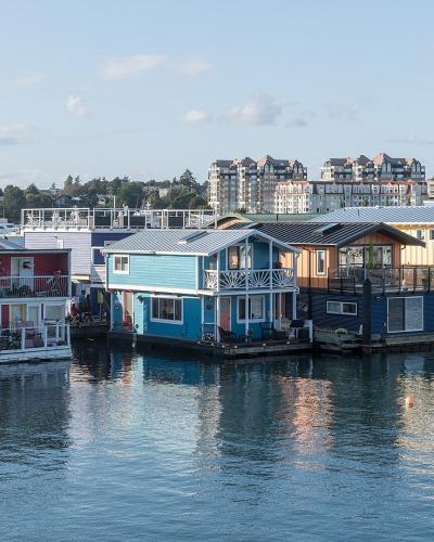 A row of colorful, multi-story houseboats moored in a harbor with modern apartment buildings visible on the shoreline in the background.