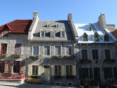 A row of historic stone buildings with slate roofs, dormer windows, and flower boxes situated on a paved square under a clear blue sky.