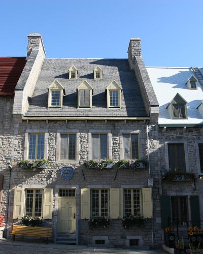 A row of historic stone buildings with slate roofs, dormer windows, and flower boxes situated on a paved square under a clear blue sky.