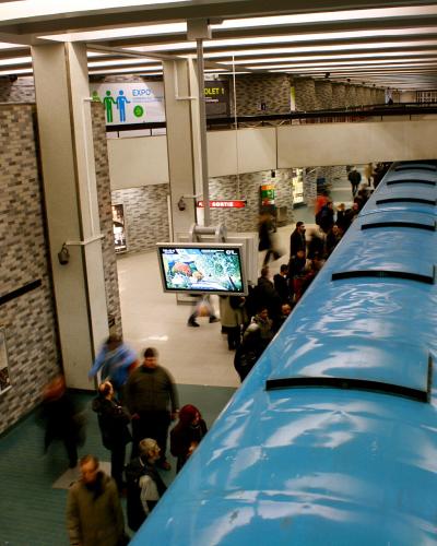 An interior view of a Montreal Metro station platform showing a blue train, tiled walls, and signage reading "PLACE-DES-ARTS".