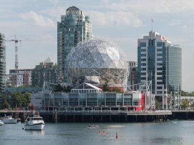 A large geodesic dome structure situated on a pier in an urban waterfront setting, with boats and kayaks in the foreground and high-rise buildings in the background.