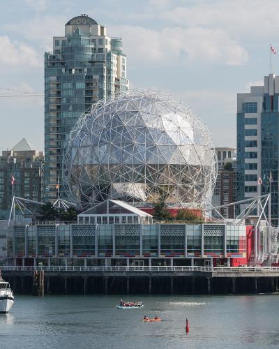 A large geodesic dome structure situated on a pier in an urban waterfront setting, with boats and kayaks in the foreground and high-rise buildings in the background.