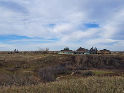 A wide landscape shot showing a modern visitor center complex with distinctive peaked roofs and a circular tower, situated on a grassy hill overlooking a valley, with a large wooden totem pole visible in the distance to the left.
