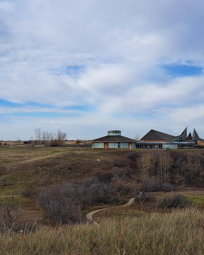 A wide landscape shot showing a modern visitor center complex with distinctive peaked roofs and a circular tower, situated on a grassy hill overlooking a valley, with a large wooden totem pole visible in the distance to the left.