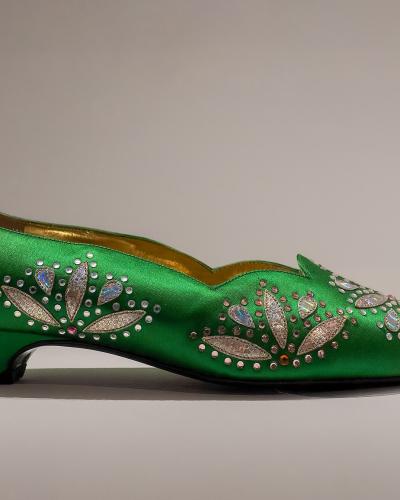 A side-profile studio photograph of an ornate green satin women's shoe decorated with silver and iridescent rhinestone floral motifs, displayed against a plain neutral background.